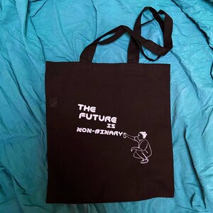 Könnte beinhalten: Schwarze Tragetasche mit weißem Text "The Future is Non-Binary" und einer weißen Illustration einer Person, die mit einer Sprühdose malt.