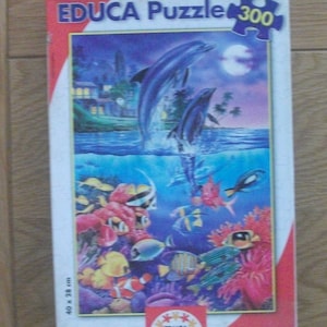Op de afbeelding: Een 300-delige puzzel met een kleurrijke onderwater scène met dolfijnen die uit het water springen, koraalriffen en tropische vissen. De doos is rood en wit met de tekst "EDUCA Puzzle 300" en een afbeelding van een puzzelstuk.