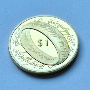 Può includere: Una moneta color oro con un design ad anello ovale e iscrizioni, contrassegnata con "$1". La moneta è posta su uno sfondo azzurro chiaro. Il design dell'anello è il fulcro centrale.
