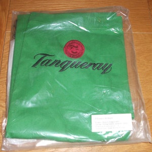 Op de afbeelding: Groene canvas draagtas met een rood en zwart geborduurd Tanqueray-logo. De tas heeft een wit label met de tekst "Klant: B14620, Bestelling: 1102713921/01, Artikel: TANQUERAY APRON, Leverancier: Adam Printer, 5/10/04".