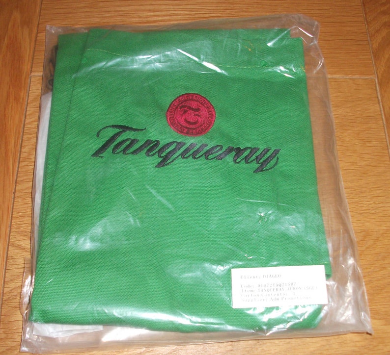 Puede incluir: Bolsa de lona verde con un logotipo Tanqueray bordado en rojo y negro. La bolsa tiene una etiqueta blanca con el texto "Cliente: B14620, Pedido: 1102713921/01, Art&iacute;culo: TANQUERAY APRON, Proveedor: Adam Printer, 5/10/04".