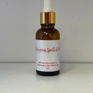 May include: A brown glass bottle with a gold lid and a white dropper top. The bottle is labeled "Obsession Spelled Oil" and "Spiritual Elite Candle Co Handmade in Fort Worth, Tx 1 oz."