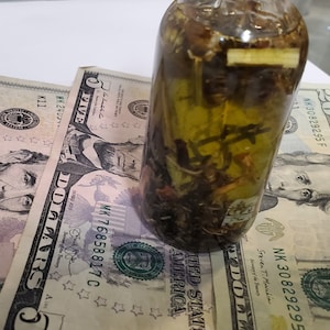 Peut inclure: Une bouteille en verre transparente remplie d'un liquide vert et de diverses herbes et &eacute;pices. La bouteille est pos&eacute;e sur cinq billets de cinq euros.