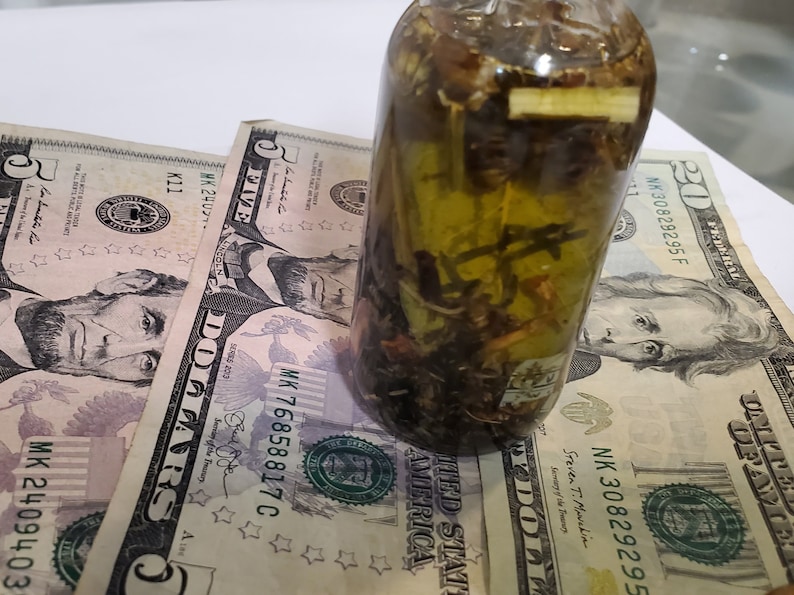 Peut inclure: Une bouteille en verre transparente remplie d'un liquide vert et de diverses herbes et &eacute;pices. La bouteille est pos&eacute;e sur cinq billets de cinq euros.