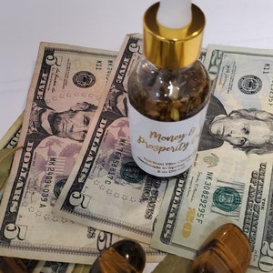 Peut inclure: Une bouteille en verre transparent avec un bouchon et un compte-gouttes dor&eacute;s remplie d'un liquide brun. La bouteille est &eacute;tiquet&eacute;e "Money & Prosperity" et est entour&eacute;e de billets de dollars am&eacute;ricains et de pierres &oelig;il de tigre.