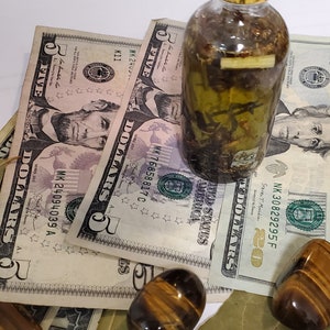 Peut inclure: Une bouteille en verre transparent avec un couvercle dor&eacute; remplie d'un liquide vert et d'herbes. La bouteille est pos&eacute;e sur des billets de cinq dollars et deux pierres &oelig;il de tigre.