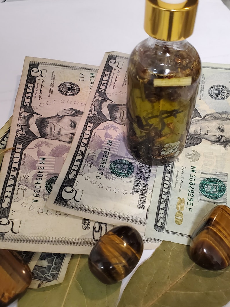 Peut inclure: Une bouteille en verre transparent avec un couvercle dor&eacute; remplie d'un liquide vert et d'herbes. La bouteille est pos&eacute;e sur des billets de cinq dollars et deux pierres &oelig;il de tigre.