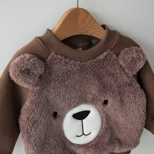 May include: A brown sweatshirt with a fuzzy brown bear face on the front. The bear has a white nose and black eyes.