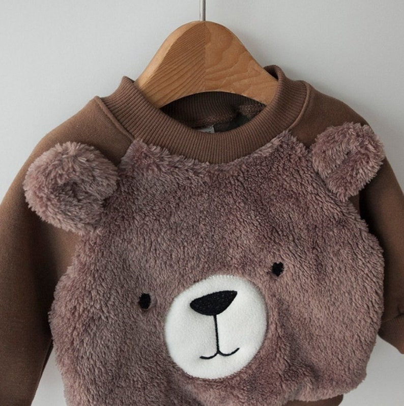 May include: A brown sweatshirt with a fuzzy brown bear face on the front. The bear has a white nose and black eyes.