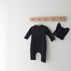 Op de afbeelding: Een zwarte baby romper en een zwarte muts hangen aan een houten rek met haken.