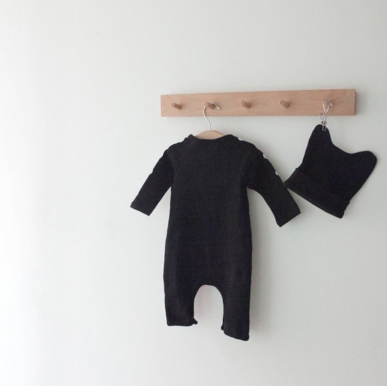 Op de afbeelding: Een zwarte baby romper en een zwarte muts hangen aan een houten rek met haken.