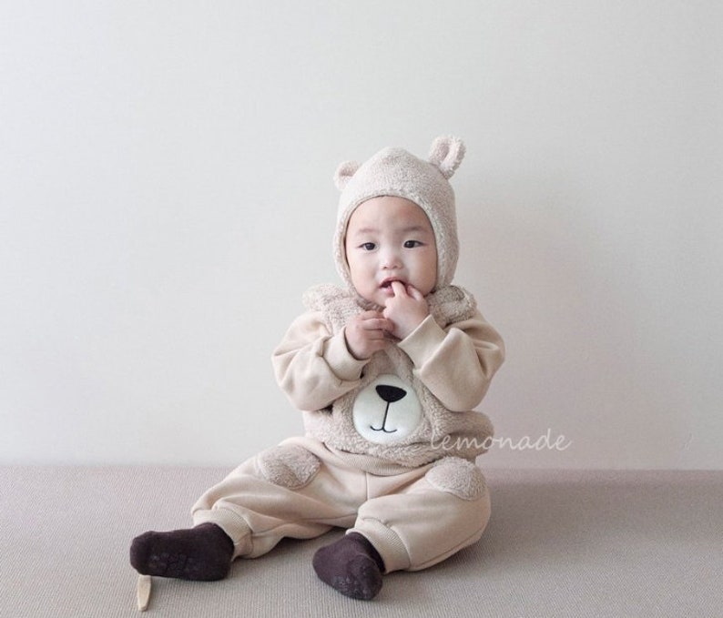 May include: A baby wearing a beige teddy bear costume with a matching hat. The costume features a brown bear face with black eyes and a black nose. The baby is sitting on a light brown surface.