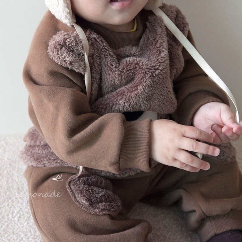 May include: A brown fleece baby outfit with a teddy bear design. The outfit has a hood with a white trim and a white ribbon tied around the neck.