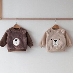 May include: Two brown and beige children's sweaters with bear faces on the front. The sweaters are hanging on hangers against a white wall.