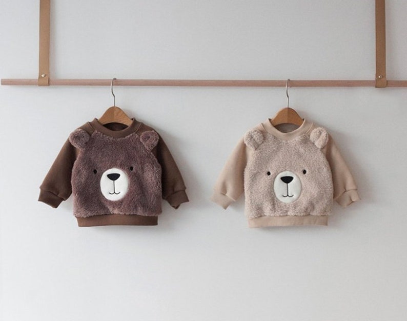 May include: Two brown and beige children's sweaters with bear faces on the front. The sweaters are hanging on hangers against a white wall.