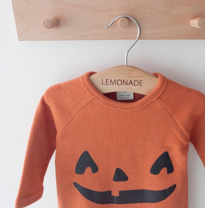 Op de afbeelding: Oranje sweatshirt met een zwart jack-o'-lantern gezicht op de voorkant. De sweatshirt hangt aan een houten hanger met het woord "LEMONADE" erop.