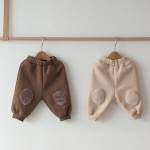 May include: Two pairs of children's sweatpants with contrasting colored fleece patches on the knees. One pair is brown and the other is beige.