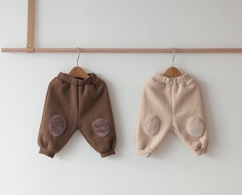 May include: Two pairs of children's sweatpants with contrasting colored fleece patches on the knees. One pair is brown and the other is beige.