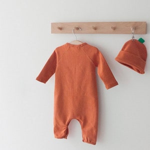 Op de afbeelding: Een oranje baby-onesie met lange mouwen en een pompoenmuts. De onesie hangt aan een houten rek met haken.