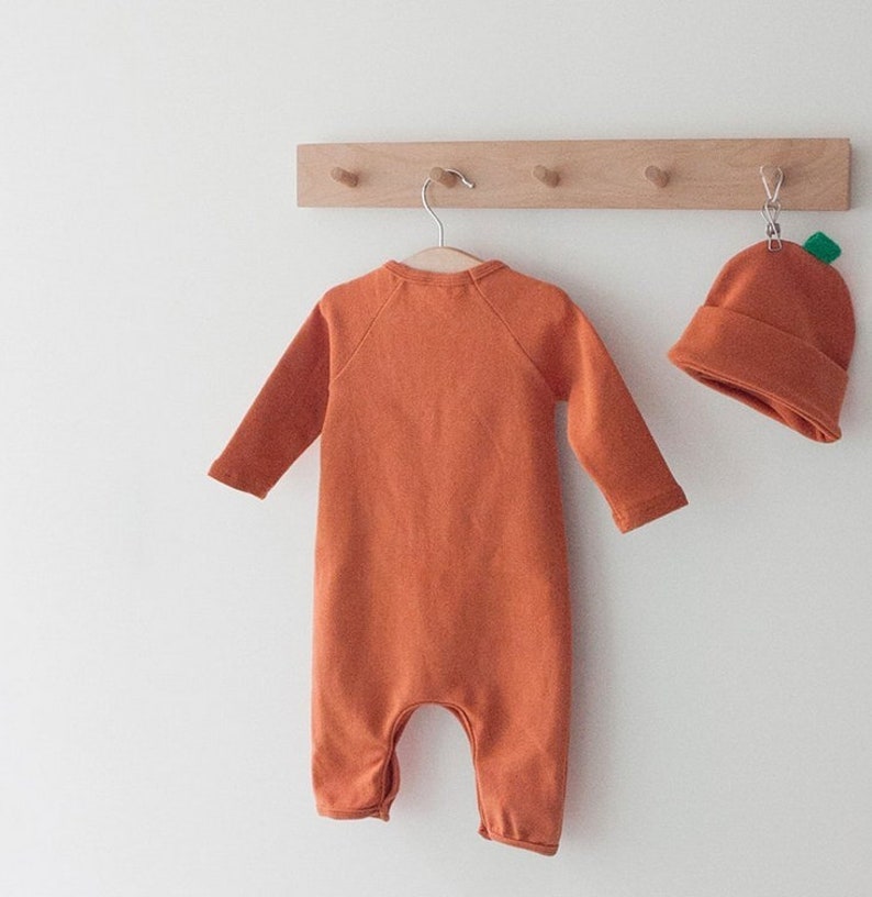 Op de afbeelding: Een oranje baby-onesie met lange mouwen en een pompoenmuts. De onesie hangt aan een houten rek met haken.