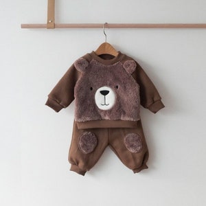 May include: Brown baby outfit with a bear face on the front of the sweatshirt and bear paw prints on the knees of the pants.