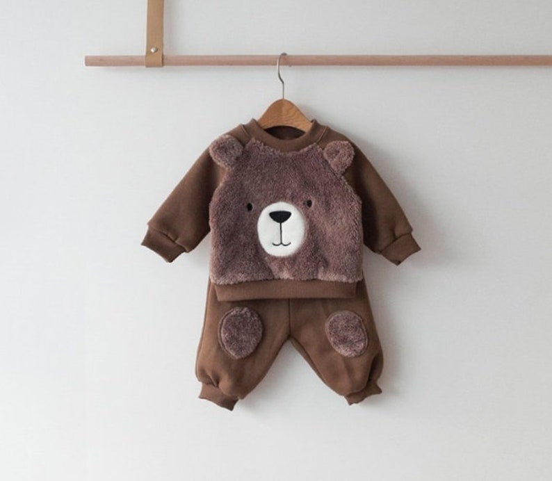 May include: Brown baby outfit with a bear face on the front of the sweatshirt and bear paw prints on the knees of the pants.
