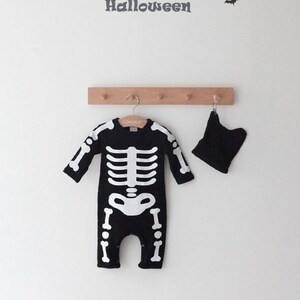 Op de afbeelding: Zwart skeletkostuum voor een baby met een bijpassende zwarte muts. Het kostuum heeft een wit skeletontwerp op de voorkant. De tekst "HAPPY HALLOWEEN" staat op een witte muur op de achtergrond.
