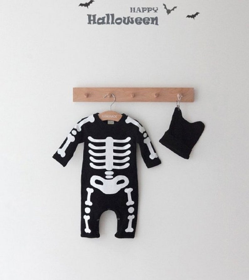 Op de afbeelding: Zwart skeletkostuum voor een baby met een bijpassende zwarte muts. Het kostuum heeft een wit skeletontwerp op de voorkant. De tekst "HAPPY HALLOWEEN" staat op een witte muur op de achtergrond.