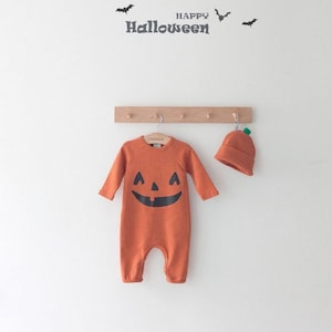 Op de afbeelding: Oranje baby-onesie met een pompoen-thema, een zwart pompoengezicht en lange mouwen. Een bijpassende oranje pompoenmuts hangt naast de onesie. De tekst "HAPPY HALLOWEEN" staat boven de onesie.