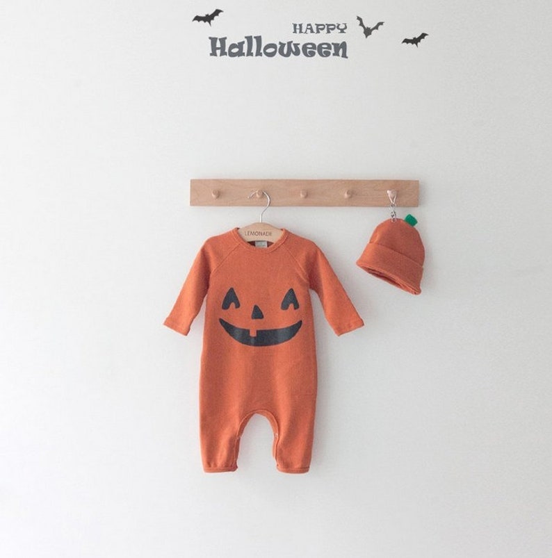 Op de afbeelding: Oranje baby-onesie met een pompoen-thema, een zwart pompoengezicht en lange mouwen. Een bijpassende oranje pompoenmuts hangt naast de onesie. De tekst "HAPPY HALLOWEEN" staat boven de onesie.