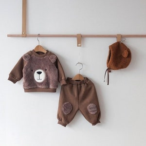 May include: A brown fleece baby outfit with a bear face on the sweater and bear paw patches on the pants. The outfit is hanging on a wooden rod with a brown fleece bear hat.
