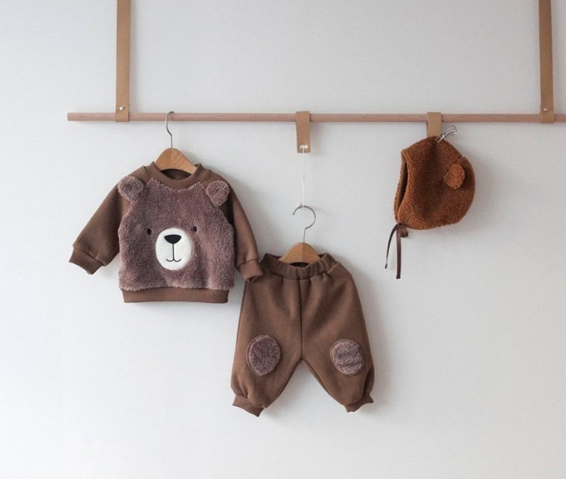 May include: A brown fleece baby outfit with a bear face on the sweater and bear paw patches on the pants. The outfit is hanging on a wooden rod with a brown fleece bear hat.