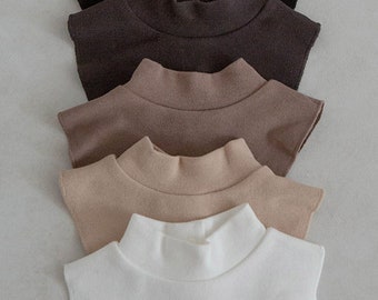 May include: A set of five mock neck collar dickies arranged in a vertical stack. The colours include black, dark brown, medium brown, beige, and white. Each piece features a high neckline and a button closure at the back of the neck for secure fastening.