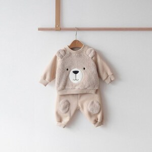 May include: A beige baby outfit with a bear face on the top and bear paw patches on the pants.