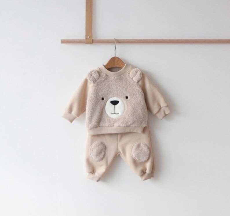 May include: A beige baby outfit with a bear face on the top and bear paw patches on the pants.