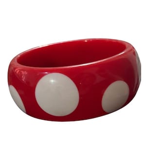May include: A thick, circular bangle bracelet featuring a vibrant red base color with large, evenly spaced white polka dots. The accessory has a smooth, glossy finish and a wide, rounded profile that creates a bold, continuous circular shape.
