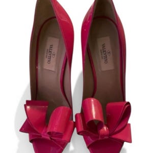 May include: A pair of bright pink patent leather high-heeled pumps featuring a large, structured bow detail on the rounded toe. The interior insole is light beige and displays the Valentino Garavani brand logo in black text on the heel area.