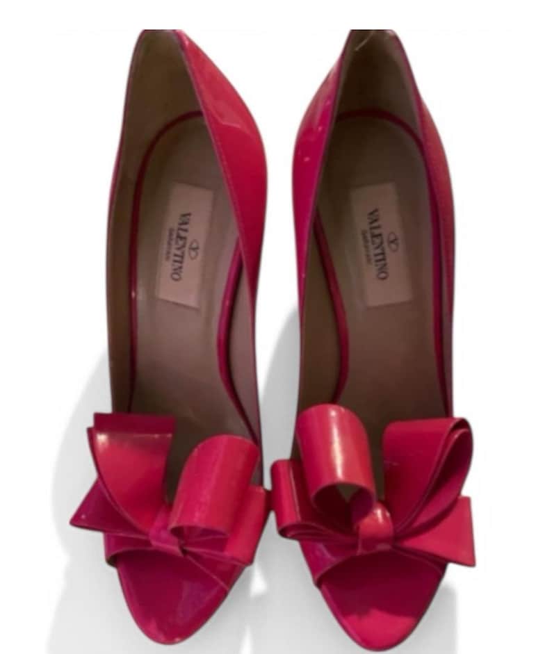 May include: A pair of bright pink patent leather high-heeled pumps featuring a large, structured bow detail on the rounded toe. The interior insole is light beige and displays the Valentino Garavani brand logo in black text on the heel area.