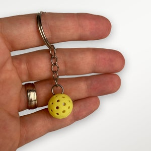 May include: A yellow, round keychain with a perforated design, resembling a pickleball. The keychain is attached to a silver chain.