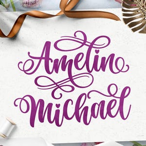 Pode incluir: Um cartão retangular branco com o texto caligráfico roxo Ameline Michael em uma fonte cursiva decorativa com pequenos corações, rodeado por uma fita marrom, flores cor-de-rosa e um elemento decorativo dourado em forma de folha.