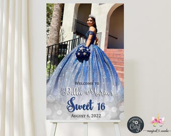 Könnte beinhalten: Ein Schild auf einer Staffelei mit der Aufschrift "Welcome to Bella Maria's Sweet 16 August 6, 2022". Es zeigt ein Foto einer Person in einem blauen Kleid mit Tiara und Blumenstrauß. Der Hintergrund ist weiß mit silbernem Bokeh.