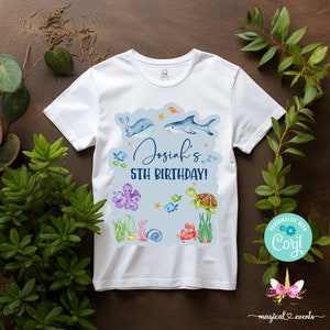 May include: White t-shirt with a blue, green, and orange ocean-themed design. The design includes a whale, a shark, an octopus, a turtle, and a crab. The text "Josiah's 5th Birthday!" is printed on the shirt.