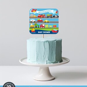 May include: A light blue frosted cake on a white cake stand, topped with a square cake topper. The topper features various modes of transportation, including a plane, cars, and a train, with the text "MEGHAN & MARK'S BABY SHOWER".