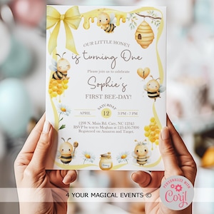 May include: A white invitation card with a bee theme. The card features illustrations of bees, honey, and daisies, with the text "Our Little Honey is turning One" and "Sophie's First Bee-Day!" The date and time are also included.