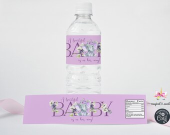 May include: A clear water bottle with a label that reads "A beautiful BABY is in her way!" in purple script. The label features a watercolor illustration of a gray elephant with floral accents. A matching label is below the bottle.