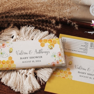 May include: A white chocolate bar wrapper with a bee and honeycomb design. The wrapper has the text "Valeria & Anthony's Baby Shower August 18, 2024".