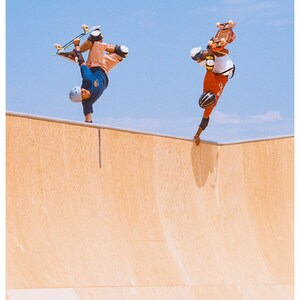 May include: Two skateboarders performing tricks in a skatepark. One skater is upside down with their board above their head, while the other skater is upside down with their board below their feet. Both skaters are wearing helmets and protective gear.