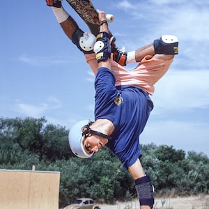 May include: A skateboarder wearing safety gear performs a handstand on a skateboard ramp. The skateboarder is wearing a blue shirt, pink shorts, a white helmet, and knee and elbow pads. The skateboarder is holding the skateboard with one hand and has the other hand on the ground. The skateboarder is in mid-air with the skateboard above their head.