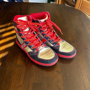May include: A pair of high-top sneakers with red laces and a gold and black design. The shoes have a red and black trim around the sole.
