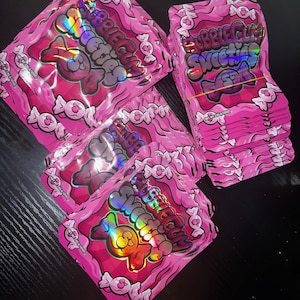 Pu&ograve; includere: Confezioni di gomma da masticare rosa con testo e grafica olografici. Le confezioni sono impilate e sparse, con le parole "Bubble Gum Sweeties" ben visibili. Il design include caramelle e un personaggio dei cartoni animati.